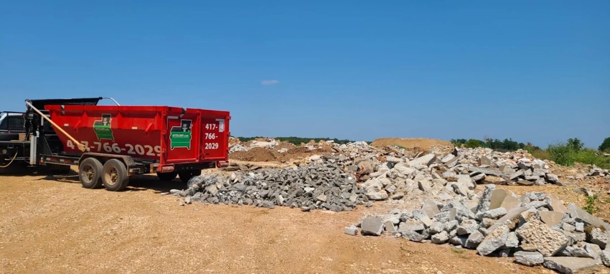 Red roll-off dumpster on a dirt site beside piles of broken concrete