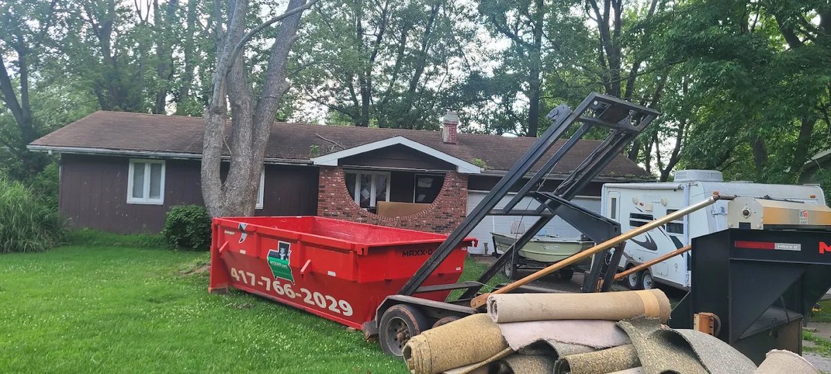 Red roll-off dumpster on a lawn with rolled-up old carpet stacked beside it