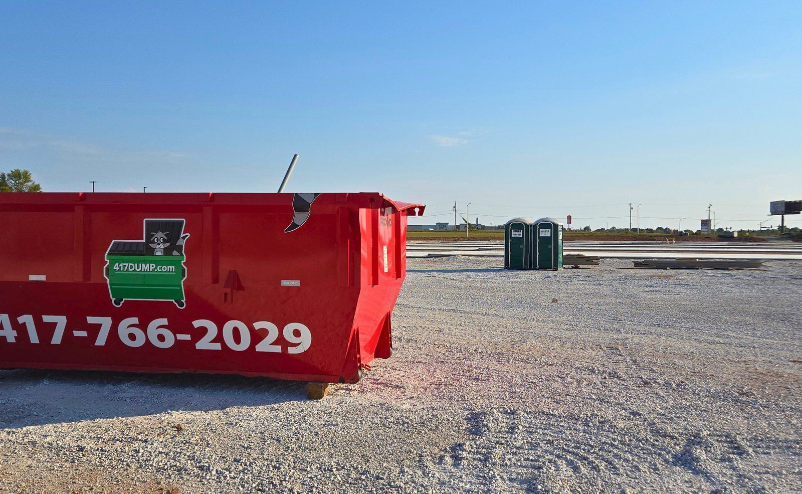 Red roll-off dumpster on a gravel job-site pad with portable toilets in the background.