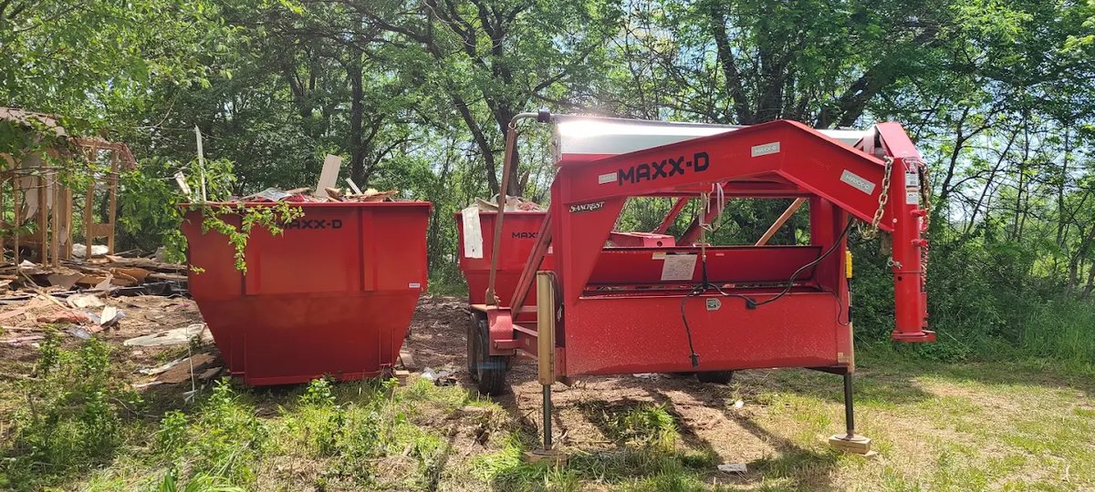Red roll-off dumpster and trailer beside a house with cut brush and tree limbs