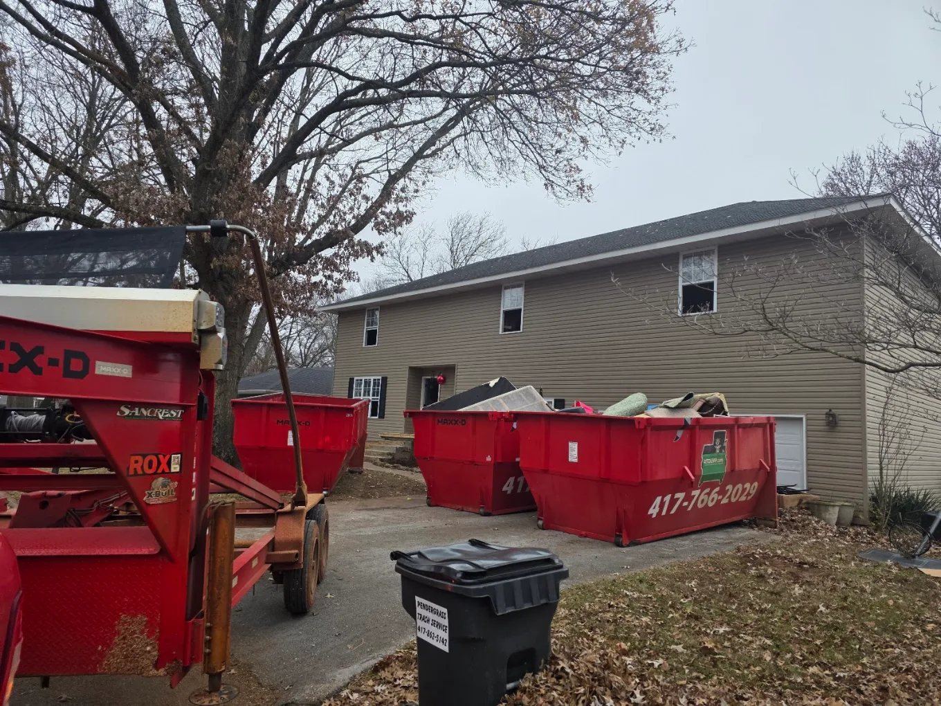 Trailer and red roll-off dumpster parked on a residential street beside a house